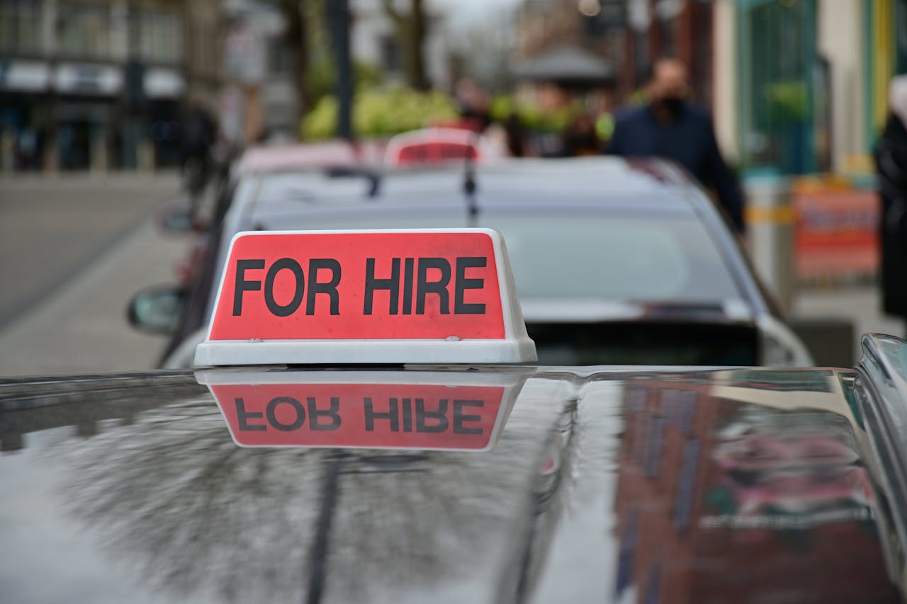 Close-up of a red 'For Hire' sign on a taxi, reflecting the urban street.
